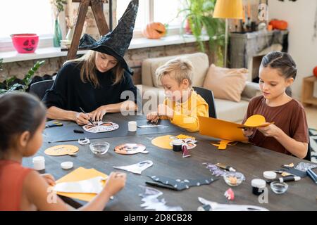 Konzentrierte junge Kunstlehrerin in Hexenhut am Tisch sitzen Und zeichnen Halloween-Bilder mit Kindern während der Vorbereitung Papierdekorationen Stockfoto