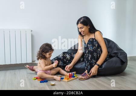 Baby Mädchen, 2 Jahre alt, spielt auf einem Boden mit ethnischen Latino Mutter Puzzle-Spielzeug, während Mutter in Blumenkleid sieht sie auf einem schwarzen Puff auf dem f sitzen Stockfoto