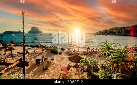 Malerischer Blick auf Cala d'Hort tropischen Strand, Menschen hängen in schönen Strand mit Es Vedra Felsblick während herrlichen lebhaften Sonnenuntergang glühenden su Stockfoto