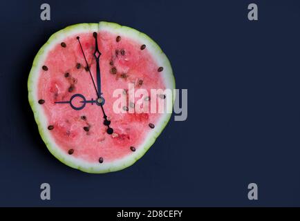 Wassermelonenscheibe mit Pfeilen, die neun Uhr auf der Uhr zeigen, dunkelblauer Hintergrund. Speicherplatz kopieren Stockfoto