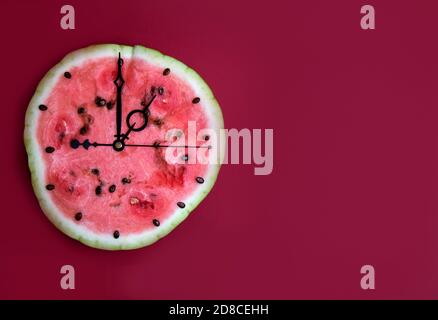 Wassermelonenscheibe mit Pfeilen, die eine Stunde auf der Uhr zeigen, roter Hintergrund. Horizontale Ausrichtung. Speicherplatz kopieren Stockfoto