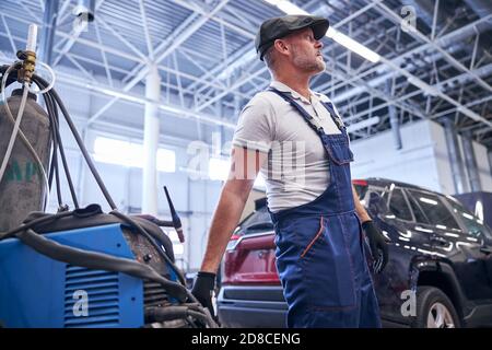 Bärtiger Mechaniker, der das fahrbare Batterieladegerät in der Autowerkstatt trägt Stockfoto