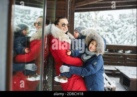 Ein Portret der schönen jungen kaukasischen Mutter mit ihren Kindern Im Winter in der Nähe des Hauses mit einem schneebedeckten weihnachten Bäume Stockfoto