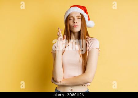 Ingwer Frau kreuzt seine Finger mit geschlossenen Augen, mit langen Haaren trägt Santa Hut mit Pouting Lippen isoliert über gelben Backgr beleidigt aussehen Stockfoto