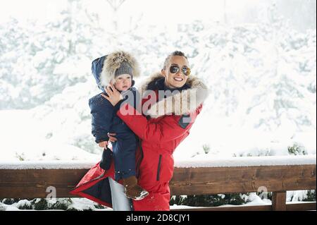 Ein Portret von schönen jungen Mutter in roter Jacke gekleidet Mit einem Kind in den Armen im Winter in der Nähe der Haus mit einem schneebedeckten weihnachtsbaum in einer BA Stockfoto