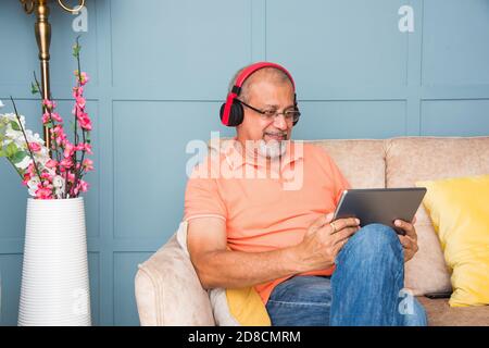 Indischer asiatischer alter Mann, der Kopfhörer benutzt, während er Musik hört Oder Filme auf einem Tablet-Computer oder Smartphone ansehen Stockfoto