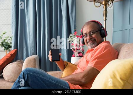 Indischer asiatischer alter Mann, der Kopfhörer benutzt, während er Musik hört Oder Filme auf einem Tablet-Computer oder Smartphone ansehen Stockfoto