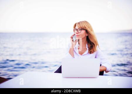 Aufnahme einer schönen reifen Frau, die mit jemandem auf ihrem Handy spricht und einen Laptop benutzt, während sie am Meer sitzt. Stockfoto