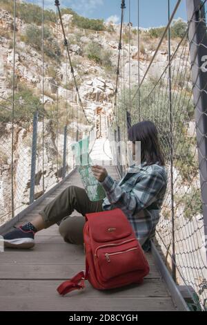 Backpacker Mädchen auf einer Hängebrücke in Chulilla sitzen, Blick auf ihre Karte, um die Gegend zu lokalisieren.Outdoor-Aktivitäten. Auf der Karte auf Hängebrücke. Stockfoto