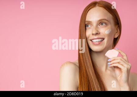 Headshot schöne fröhliche rothaarige Frau mit Sauber Gesunde Haut, das Anwenden der Grundlage mit kosmetischen Schwamm, lächelnd weiße Zähne auf isolierte Stockfoto