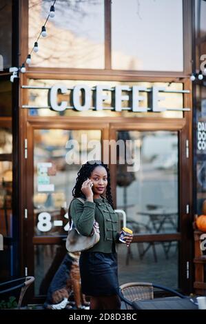 Stilvolle modische afroamerikanische Frauen in grünen Pullover und schwarzen Rock posiert Outdoor-Café mit Tasse Kaffee und sprechen auf Handy. Stockfoto