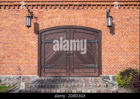 Alte Holztore mit Eisenschlaufen, Granitsteinstufen und Lampen an den Seiten in der roten Ziegelwand, aus der Nähe Stockfoto