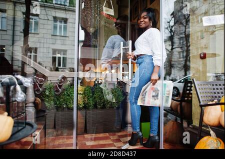 Stylische afroamerikanische Frauen in weißer Bluse und blauer Jeans posierten in der Nähe von Café mit Zeitung. Stockfoto