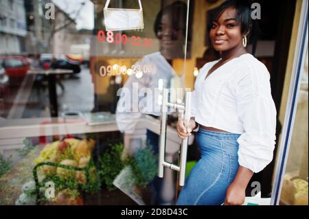 Stylische afroamerikanische Frauen in weißer Bluse und blauer Jeans posierten in der Nähe von Café mit Zeitung. Stockfoto