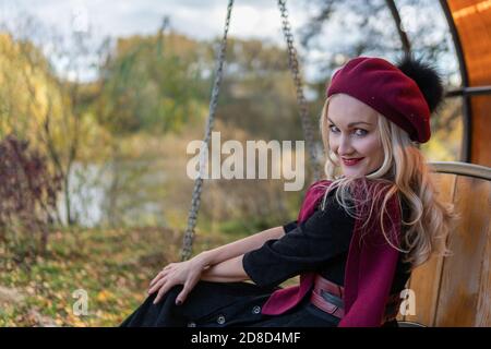 Ein Mädchen sitzt auf einer Gartenschaukel, geschmiedet in einem burgunderroten Mantel und biret, mit einem charismatischen Aussehen, im Herbst vor dem Hintergrund der Bäume die Stockfoto