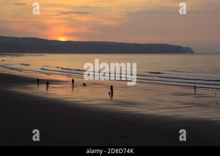 Silhouetten von Menschen, die am Whitby Beach spazieren, während die Sonne über der Ostküste untergeht, Whitby, North Yorkshire, England, Großbritannien. Stockfoto