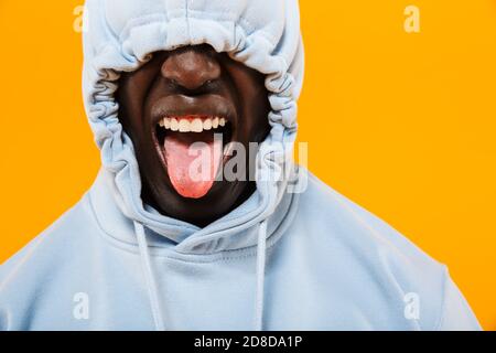 Bild von lustigen afroamerikanischen Kerl in Streetwear Hoodie kleben Aus seiner Zunge isoliert auf gelbem Hintergrund Stockfoto