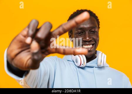 Bild von jungen afroamerikanischen Kerl in Kopfhörer lächelnd und Zeigt Friedenszeichen isoliert auf gelbem Hintergrund Stockfoto