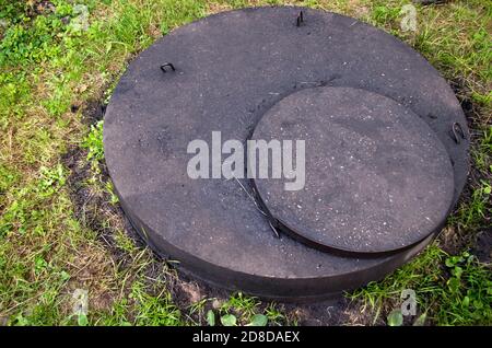 Betonbrunnen, wie das Gesicht eines Nilpferdes von oben, mit einer abgerundeten Platte darauf und einem runden Deckel mit schwarzem Bitumen bedeckt, zwischen grünem Gras Stockfoto