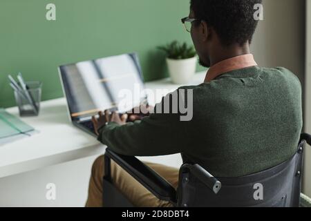 Rückansicht des afrikanischen Mannes, der auf dem Rollstuhl sitzt und auf dem tippt Laptop am Tisch im Büro Stockfoto