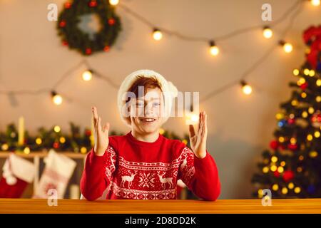 Fröhlicher Junge in Santa Hut zeigt etwas mit Händen und Blick auf die Kamera während eines Online-Meetings Stockfoto