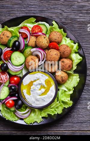 Mediterrane Platte mit Falafels, Tomaten, Gurken, roten Zwiebeln, schwarzen Oliven, auf den frischen Salatblättern serviert mit Tsatziki-Sauce auf einem schwarzen serviert Stockfoto