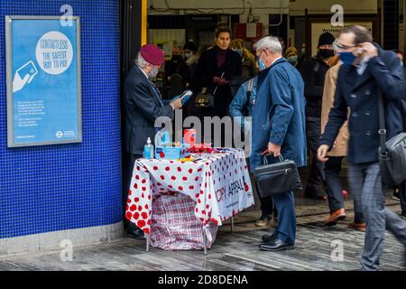 London, Großbritannien. Okt. 2020. Es ist der London Poppy Day und ein Veteran (in der Gruppe der Risikogruppen) der Luftlandetruppen, in seiner roten Baskenmütze, sammelt sich für die Royal British Legion, als ein stetiger Passagierstrom die U-Bahnstation Sloane Square verlässt. Er trägt eine Maske und Handschuhe und sammelt meist Geld mit kontaktlosen Zahlungen (obwohl er immer noch eine "Dose" für Bargeld hat). Reisende tragen meist Masken, nachdem sie obligatorisch geworden sind. Kredit: Guy Bell/Alamy Live Nachrichten Stockfoto