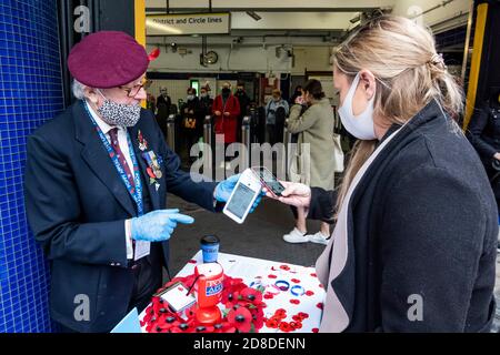 London, Großbritannien. Okt. 2020. Es ist der London Poppy Day und ein Veteran (in der Gruppe der Risikogruppen) der Luftlandetruppen, in seiner roten Baskenmütze, sammelt sich für die Royal British Legion, als ein stetiger Passagierstrom die U-Bahnstation Sloane Square verlässt. Er trägt eine Maske und Handschuhe und sammelt meist Geld mit kontaktlosen Zahlungen (obwohl er immer noch eine "Dose" für Bargeld hat). Reisende tragen meist Masken, nachdem sie obligatorisch geworden sind. Kredit: Guy Bell/Alamy Live Nachrichten Stockfoto