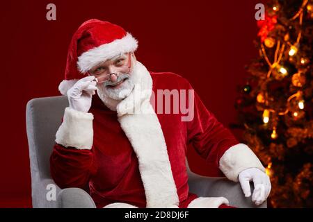 Porträt des glücklichen bärtigen Weihnachtsmann in rotem Anzug sitzend Auf dem Sessel und lächelnd in der Kamera Stockfoto