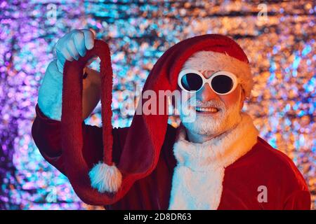 Portrait von älteren Mann in Santa Kostüm trägt Sonnenbrille und Lustige Hut lächelt auf Kamera vor dem glänzenden Hintergrund Stockfoto