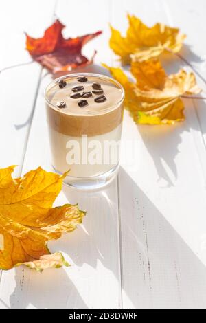 Traditionelles koreanisches koffeinhaltiges Milchgetränk, Dalgona-Kaffee in einem transparenten Glas mit Kaffeebohnen auf weißem Hintergrund. Trockene Ahornblätter. Autom Stockfoto