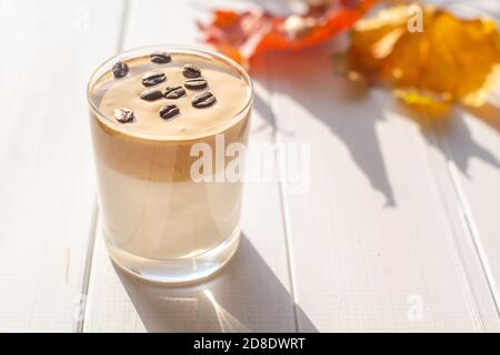 Traditionelles koreanisches koffeinhaltiges Milchgetränk, Dalgona-Kaffee in einem Glas mit Kaffeebohnen auf weißem Hintergrund. Trockene Ahornblätter. Herbstfrühstück Stockfoto