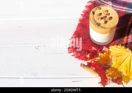 Traditionelles koreanisches koffeinhaltiges Milchgetränk, Dalgona-Kaffee in einem Glas mit Kaffeebohnen auf weißem Hintergrund. Trockene Ahornblätter. Warm karierte s Stockfoto