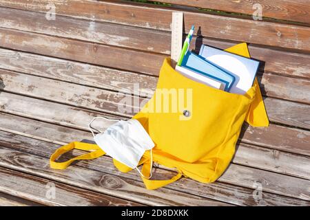 Gelber Textilrucksack mit Schulbedarf und Büchern auf einer Bank im Freien. Medizinische Maske, Virenschutz. Neues Schuljahr, zurück zur Schule, Pause. Ne Stockfoto