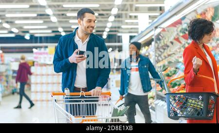 Im Supermarkt: Handsome man mit Smartphone, schiebt den Warenkorb, geht durch die Frischwarenabteilung des Stores, wählt einige Produkte. Andere Stockfoto