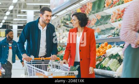 Im Supermarkt: Das glückliche junge Paar wählt Bio-Gemüse in der Frischwarenabteilung des Stores. Freund schiebt Warenkorb während Stockfoto
