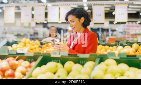 Im Supermarkt: Porträt der schönen Frau mit Smartphone, wählt Produkte im Frischegang. Frau in Internet-Surfen eingetaucht Stockfoto