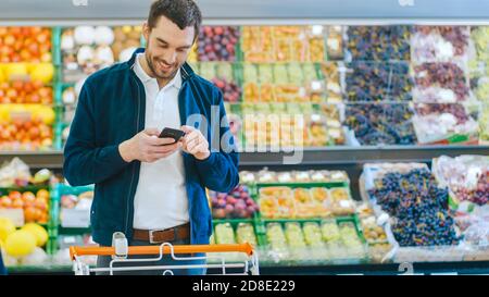 Im Supermarkt: Handsome man benutzt Smartphone, während er in der Frischwarenabteilung des Stores steht. Mann in Internet-Surfen auf seinem eingetaucht Stockfoto