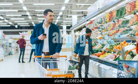 Im Supermarkt: Handsome man mit Smartphone, schiebt den Warenkorb, geht durch die Frischwarenabteilung des Stores, wählt einige Produkte. Andere Stockfoto