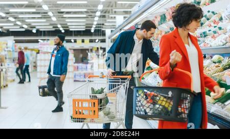 Im Supermarkt: Handsome man mit Smartphone, schiebt den Warenkorb, geht durch die Frischwarenabteilung des Stores, wählt einige Produkte. Andere Stockfoto