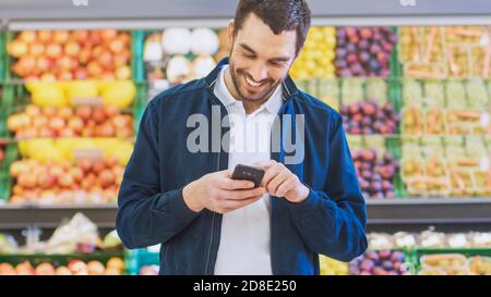 Im Supermarkt: Handsome man benutzt Smartphone, während er in der Frischwarenabteilung des Stores steht. Mann in Internet-Surfen auf seinem eingetaucht Stockfoto