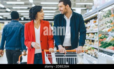 Im Supermarkt: Fröhliches junges Paar geht durch die Frischeabteilung des Stores, sie reden, Mann umarmt Frau liebevoll. Stockfoto
