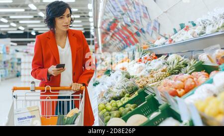 Im Supermarkt: Die schöne junge Frau geht durch die Frischeabteilung, wählt Gemüse und schiebt den Warenkorb. Kunde Nutzt Smartphone Stockfoto