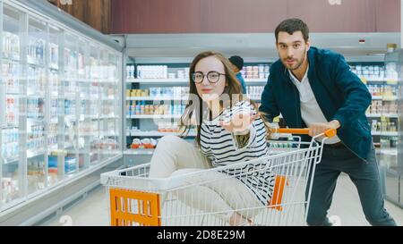 Im Supermarkt: Mann schiebt Einkaufswagen mit Frau sitzt darin, Happy Couple hat Spaß beim Racing Drift auf einem Trolley durch den Laden. Stockfoto