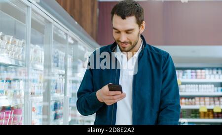 Im Supermarkt: Handsome man nutzt Smartphone, smiles und geht durch Frozen Goods Section. Andere Kunden, die im Hintergrund einkaufen. Stockfoto