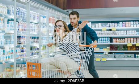 Im Supermarkt: Mann schiebt Einkaufswagen mit Frau sitzt darin, Happy Couple hat Spaß beim Rennen auf einem Trolley durch den Laden. Stockfoto