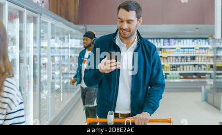 Im Supermarkt: Handsome man with Shopping Cart nutzt Smartphone, smiles und geht durch die Gefriergut-Sektion. Andere Kunden, die einkaufen Stockfoto