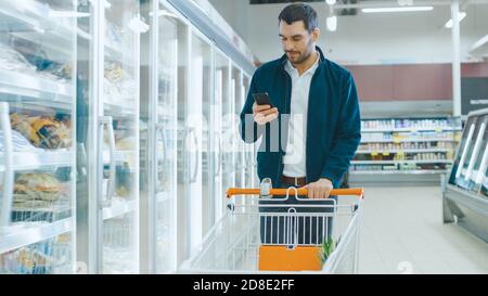 Im Supermarkt: Handsome man nutzt Smartphone, smiles und geht durch Frozen Goods Section. Andere Kunden, die im Hintergrund einkaufen. Stockfoto