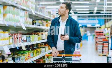 Im Supermarkt: Handsome man durchstösst Regal mit Konserven, platziert Blechdosen in seinen Warenkorb und geht mit seiner Einkaufsliste weiter Stockfoto