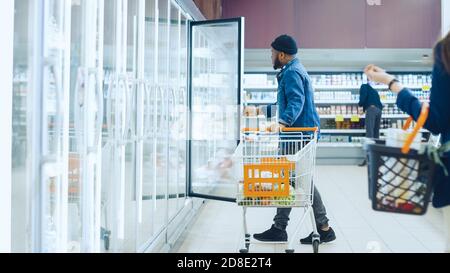 Im Supermarkt: Happy Stylish Guy schiebt den Warenkorb und wählt in der Tiefkühlwarenabteilung des Stores gefrorenes Gemüse. Große Mall mit Glas Stockfoto